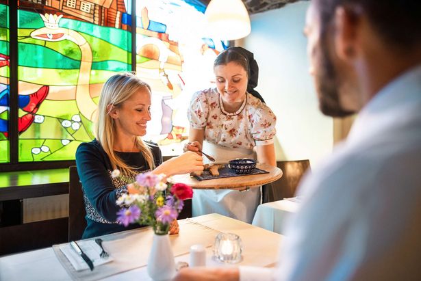 Eine Kellnerin im sorbischen Restaurant hat Brotstücke und einen Aufstrich auf einem Brettchen stehen. Eine Frau kostet gerade etwas davon. 