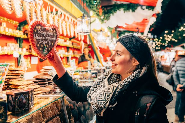 A young woman is standing in front of a gingerbread stall holding a gingerbread heart with "Greetings from Christmas Market Leipzig" written on it. 