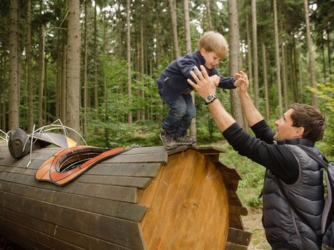 Ein Junge steht auf einem Rohr aus Holz und sein Vater hilft ihm beim Runterklettern. Er ist in der Waldhusche in der Sächsischen Schweiz.